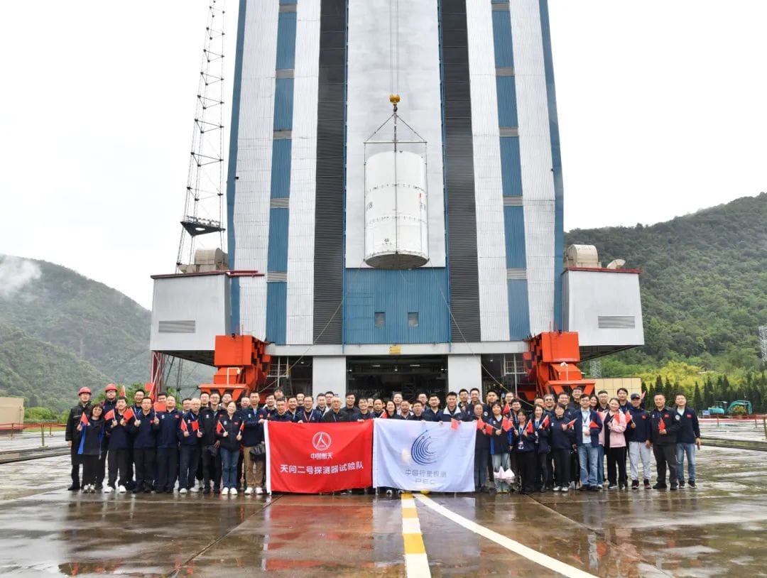 Part of the team for the Tianwen-2 mission pose with the spacecraft, in its protective container, during its lift toward the top of its Long March 3B/E rocket.
